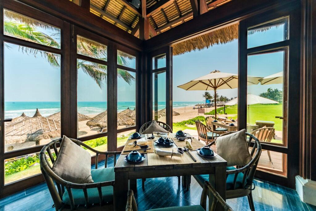 Ein Esstisch für vier Personen in einem Restaurant mit offenen Fenstern und Blick auf einen Strand mit Palmen und strohgedeckten Sonnenschirmen im luxuriösen „The Anam Cam Ranh“ in Vietnam