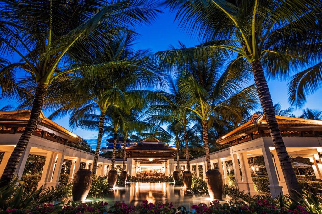 Die Open-Air-Lobby des Resorts „The Anam Cam Ranh“ mit hohen Palmen, die einen Spiegelteich säumen, und beleuchteten Sitzbereichen in der Abenddämmerung