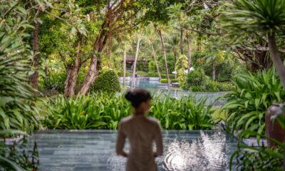 Blick über mehrere Wasserbecken und üppige Vegetation im vietnamesischen Resort „The Anam Cam Ranh“ in Richtung eines Weges; im Vordergrund steht eine Person (unscharf), die in den Hintergrund blickt