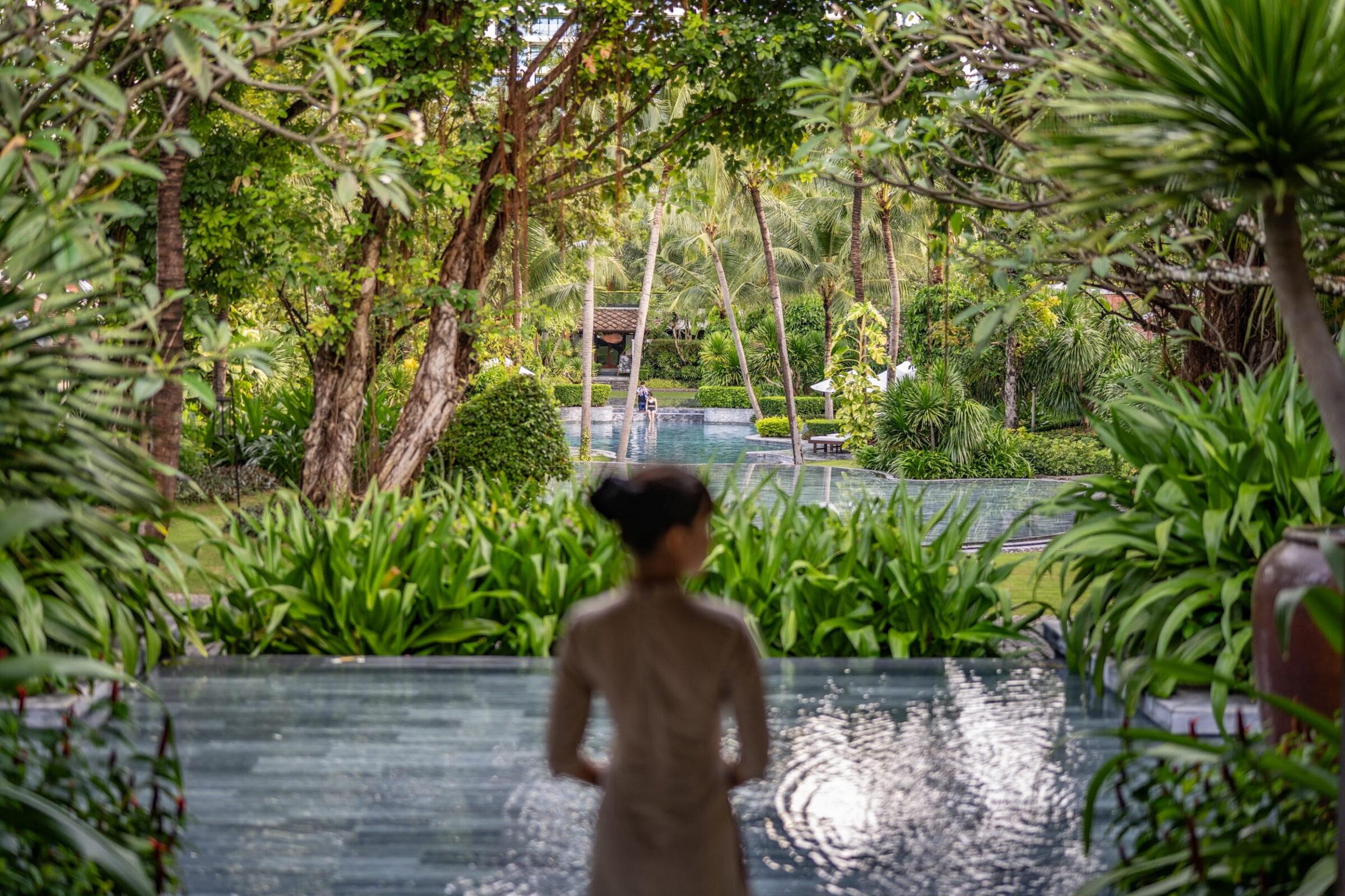 Blick über mehrere Wasserbecken und üppige Vegetation im vietnamesischen Resort „The Anam Cam Ranh“ in Richtung eines Weges; im Vordergrund steht eine Person (unscharf), die in den Hintergrund blickt