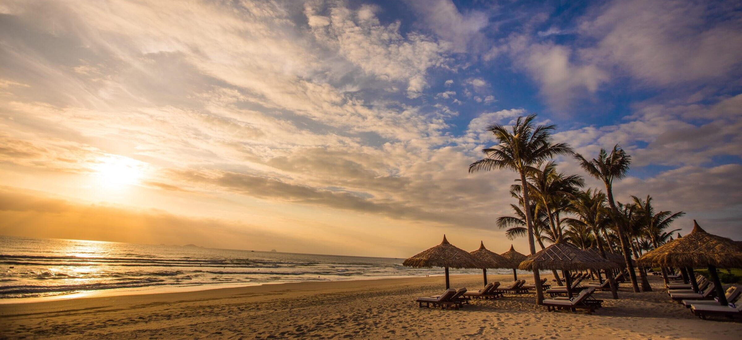 Strand mit Liegestühlen und strohgedeckten Sonnenschirmen unter Palmen bei Sonnenaufgang im Resort „The Anam Cam Ranh“ in Vietnam