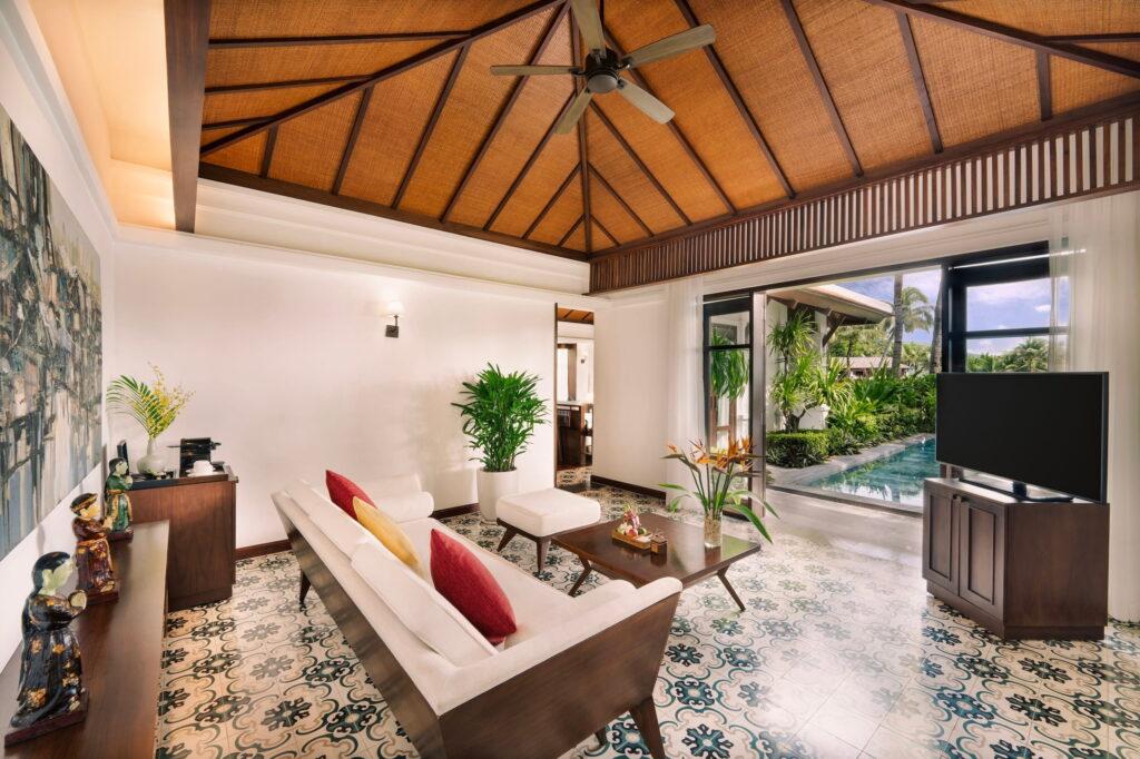 Wohnbereich einer luxuriösen Villa im Resort „The Anam Cam Ranh“ in Vietnam mit gemustertem Fliesenboden, Holzdecke mit Deckenventilator, weißem Sofa mit roten Kissen, Glas-Couchtisch, Fernseher auf einem Holzständer und Glasschiebetüren, die zum Pool und in den Garten führen