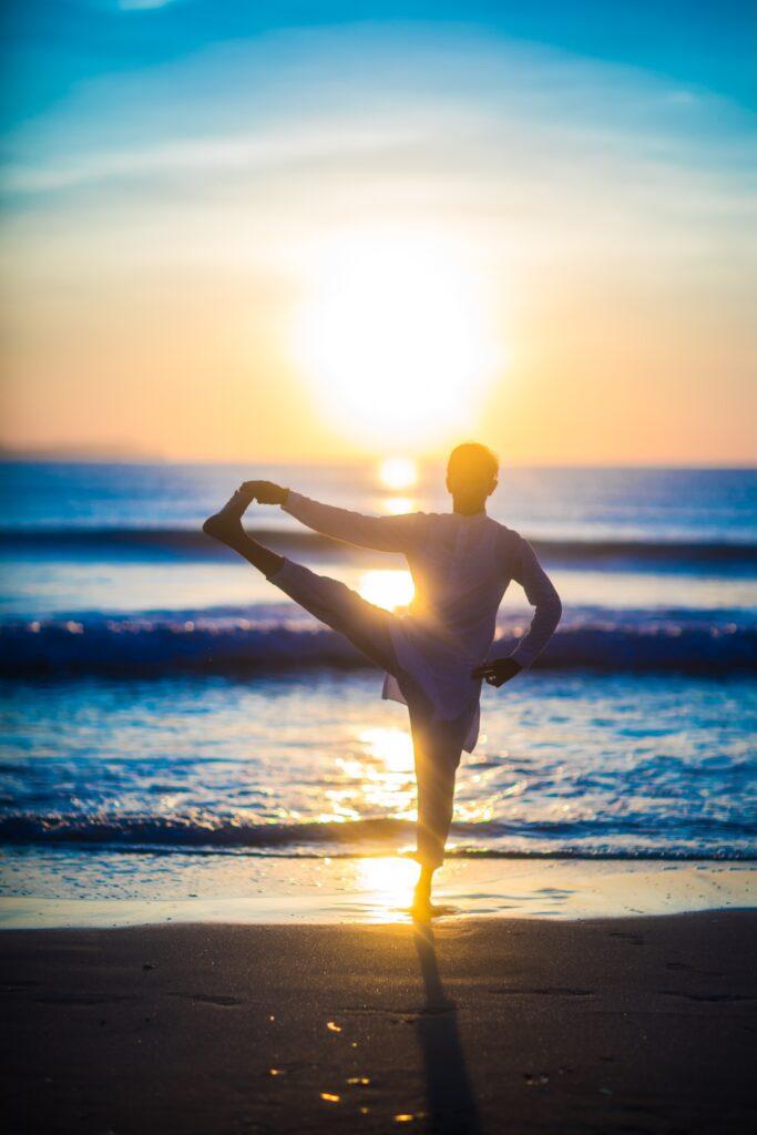 Jemand, der Yoga macht und dabei bei Sonnenaufgang am Strand auf einem Bein balanciert, während die Sonne tief über dem Horizont des Meeres steht