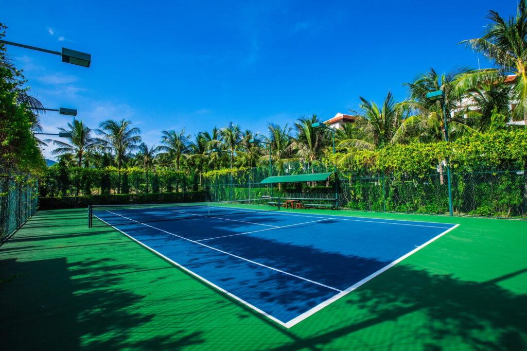 Tennisplatz im Freien mit blauem Belag und grüner Umgebung im Resort „The Anam Cam Ranh“, mit Palmen und strahlend blauem Himmel