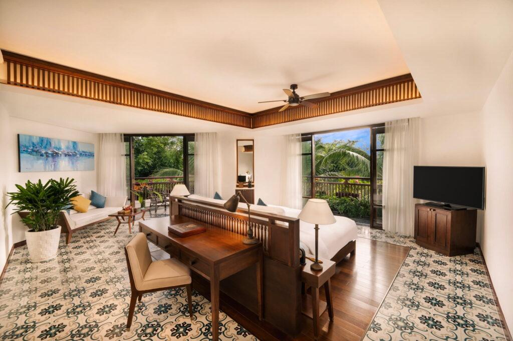 Luxuriöse Suite im „The Anam Cam Ranh“ mit zwei Betten, die durch eine Holztrennwand voneinander getrennt sind, einem Schreibtisch mit Stuhl, einem gemusterten Fliesenboden, einem Deckenventilator und großen Fenstern, die auf eine Terrasse mit viel Grün im Außenbereich führen
