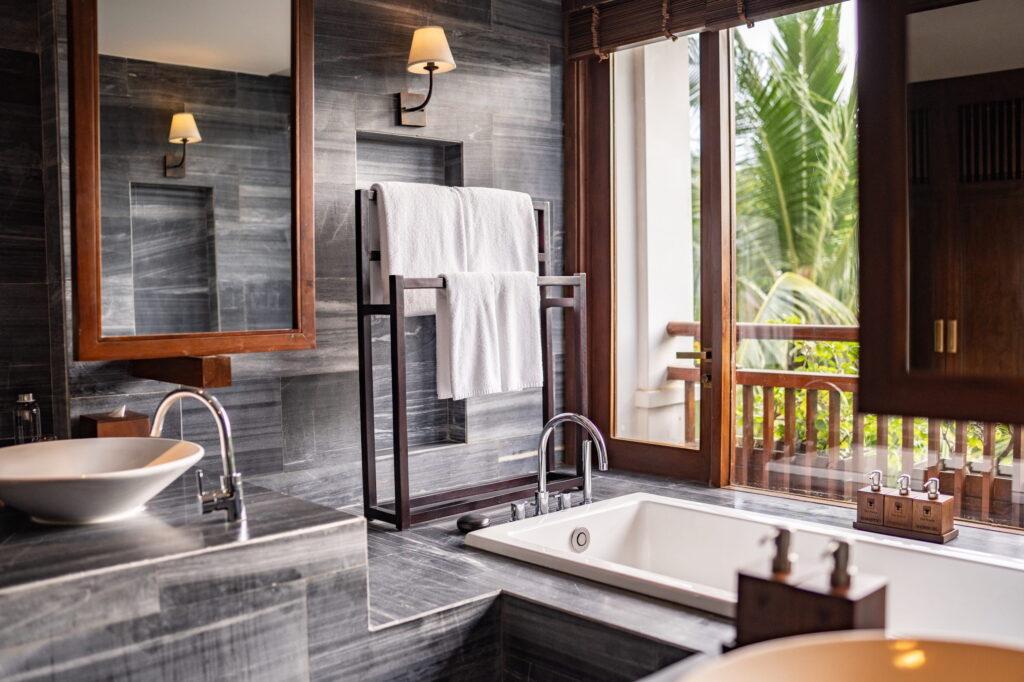 Badezimmer in einer privaten Villa im luxuriösen Resort „The Anam Cam Ranh“ mit Marmoroberflächen, einer Badewanne neben einem Fenster mit Blick auf Palmen, einem Handtuchhalter mit weißen Handtüchern und einer Wandlampe über dem Spiegel