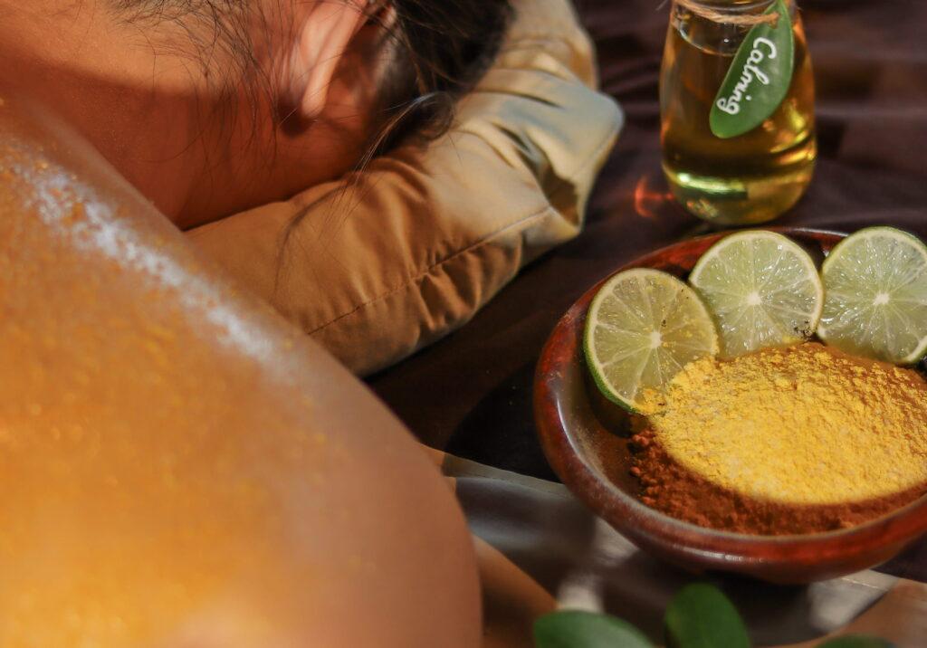 Der Rücken einer Person, die auf einer Wellnessliege entspannt, daneben eine Schale mit gelbem Pulver, Limettenscheiben und eine Flasche mit grünem Etikett