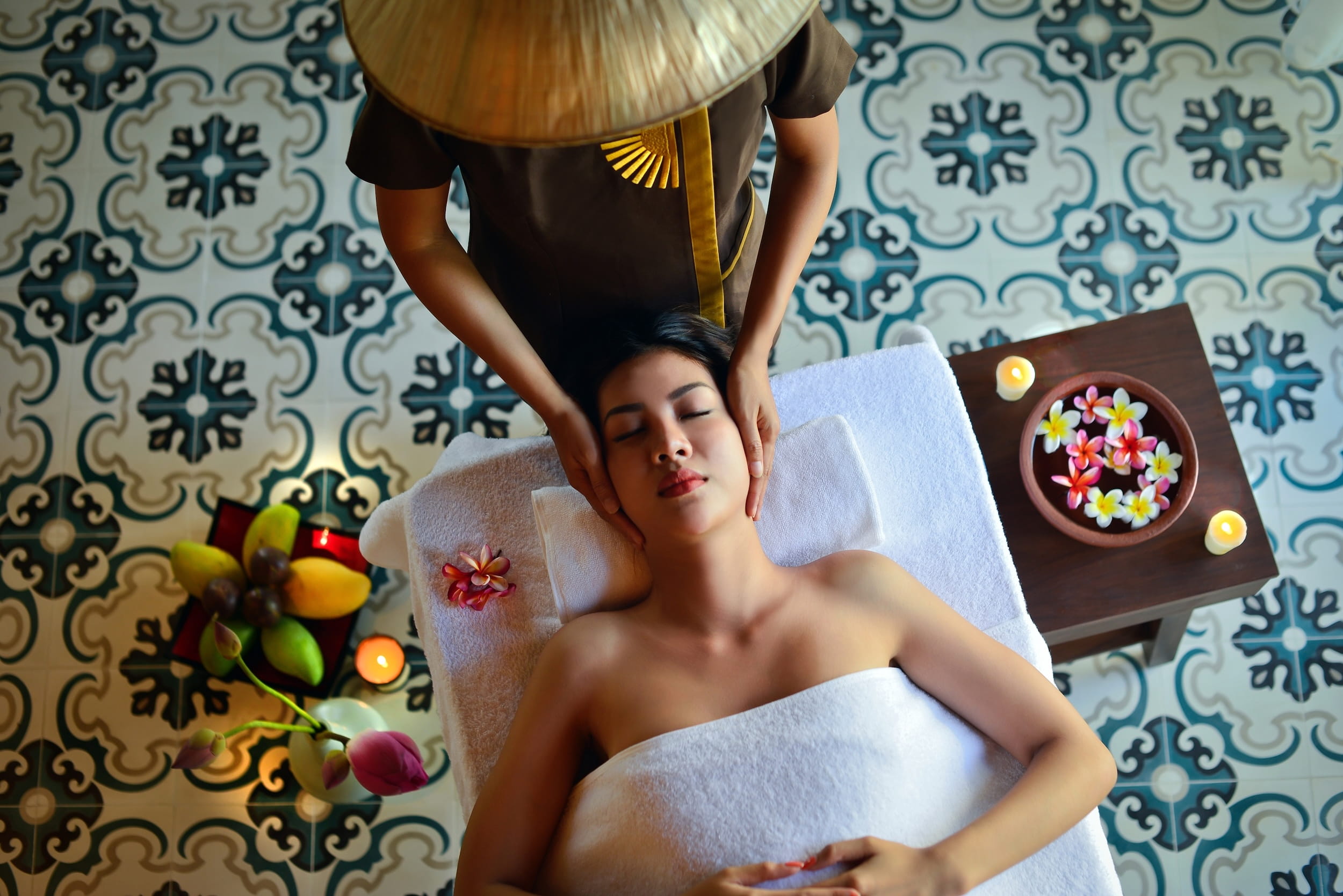 Eine Person liegt auf einer mit einem weißen Handtuch bedeckten Massageliege und wird von einer stehenden Person massiert, die einen traditionellen vietnamesischen Kegelhut trägt. Um sie herum stehen Kerzen und Blumen auf einem gemusterten Boden im The Anam Spa.