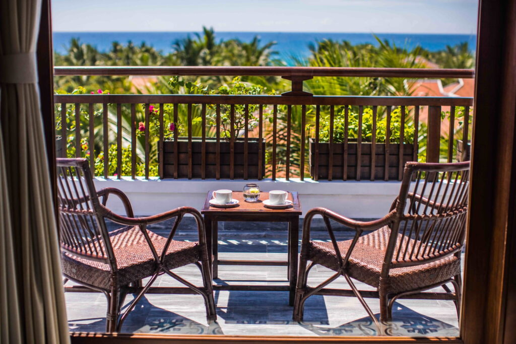 Zwei Korbstühle und ein kleiner Tisch mit zwei Tassen auf einem Balkon mit Blick ins Grüne und aufs Meer