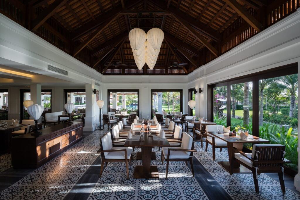 Geräumiger Restaurantbereich im Anam Cam Ranh in Vietnam mit Holztischen und -stühlen, gemustertem Fliesenboden, großen Fenstern mit Blick auf den Garten und hängenden Laternen unter einer hohen Holzdecke