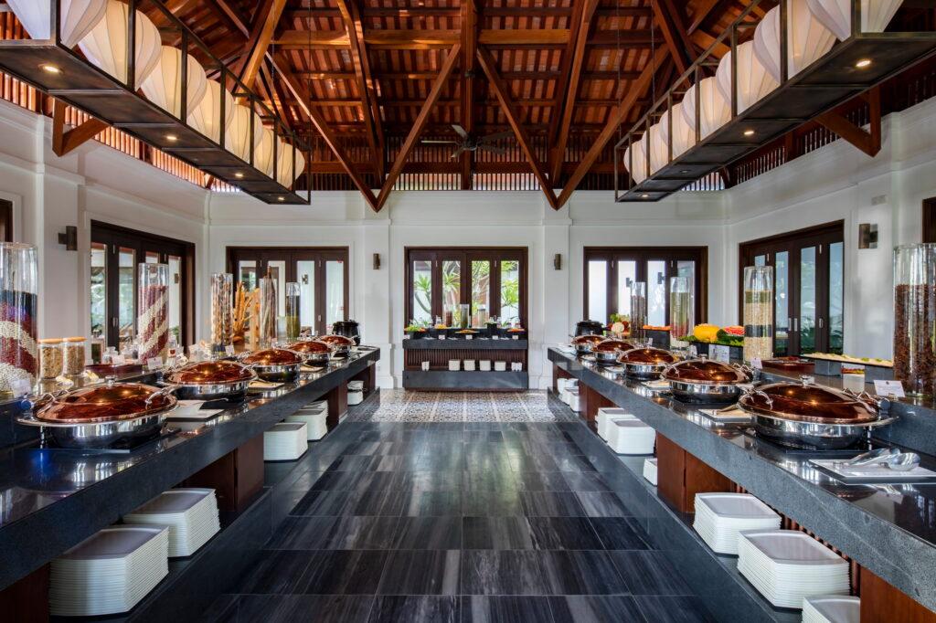 Der Innenraum des Restaurants „The Indochine“ im „The Anam Cam Ranh“ mit symmetrisch angeordneten Buffettischen und abgedeckten Speisen unter einer hohen Holzdecke mit sichtbaren Balken
