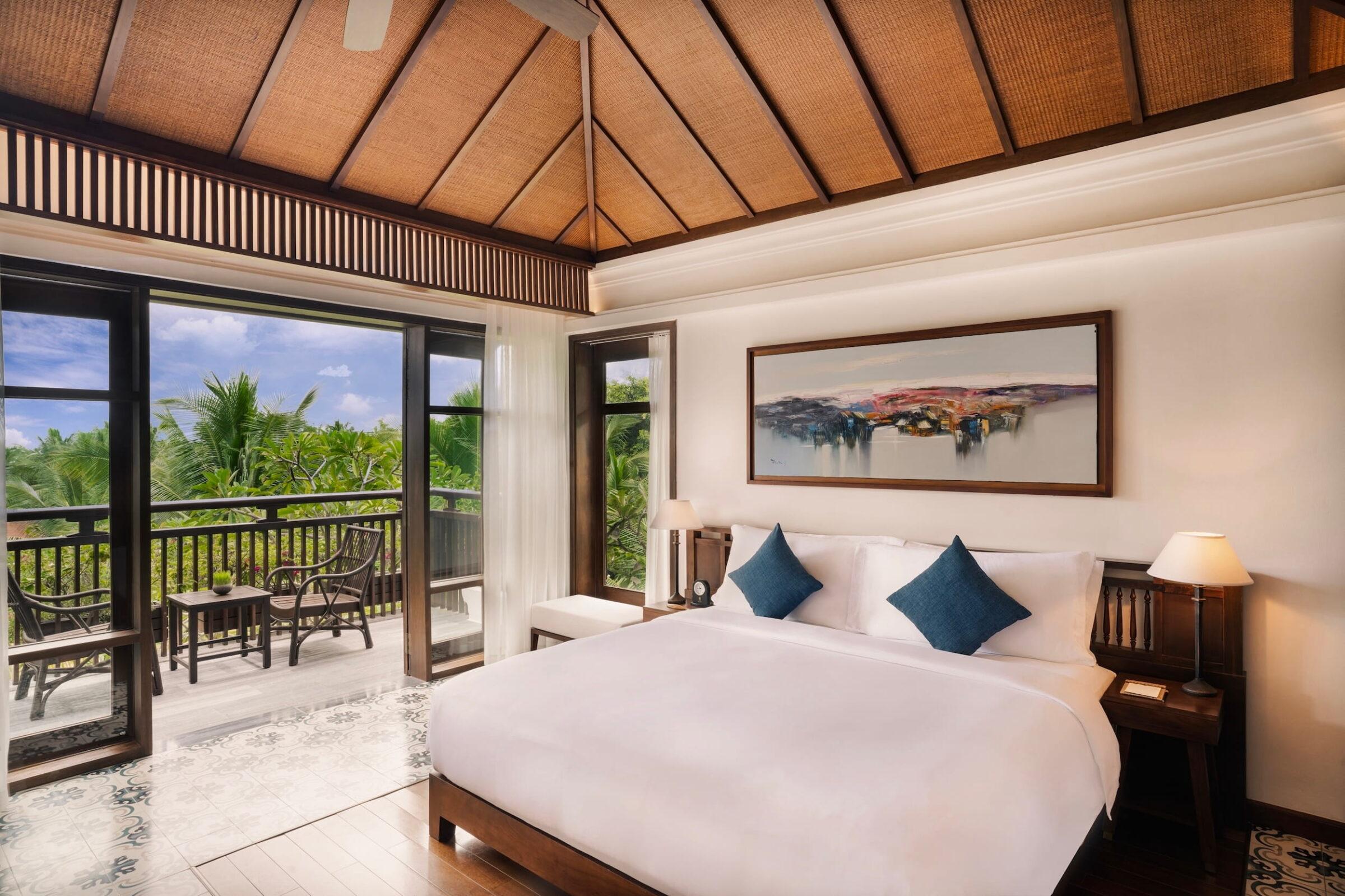 Schlafzimmer mit großem Bett, zwei blauen Kissen, Nachttischen mit Lampen, Wanddekoration und einem Balkon mit zwei Stühlen und einem Tisch in einer Villa des luxuriösen vietnamesischen Resorts „The Anam Cam Ranh“