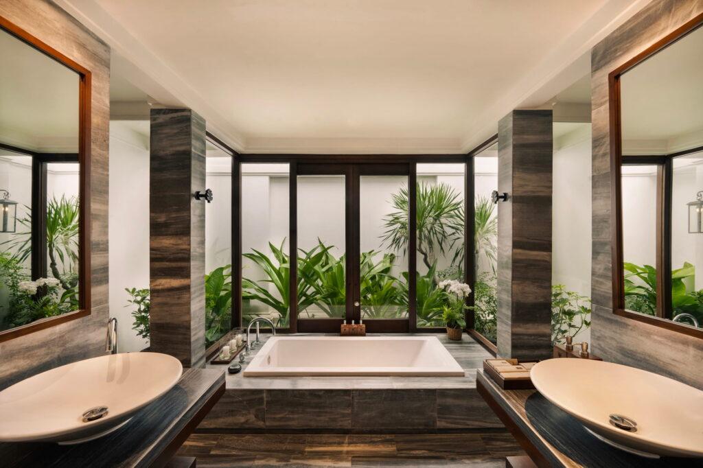 Badezimmer mit einer zentral platzierten Badewanne, flankiert von zwei ovalen Waschbecken auf dunklen Waschtischen und großen Fenstern, durch die man die grünen Pflanzen draußen sehen kann