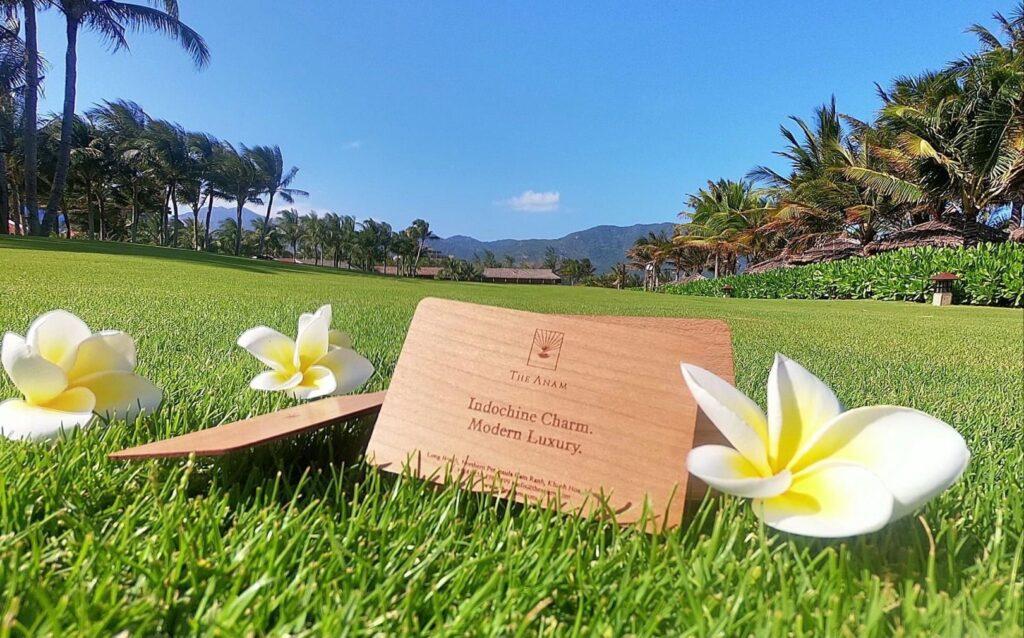 Schlüsselkarten aus Holz mit dem „The Anam“-Logo und dem Schriftzug „Indochine Charm. Modern Luxury.“ auf einer Wiese, umgeben von weißen und gelben Plumeria-Blüten, mit Palmen und Bergen im Hintergrund