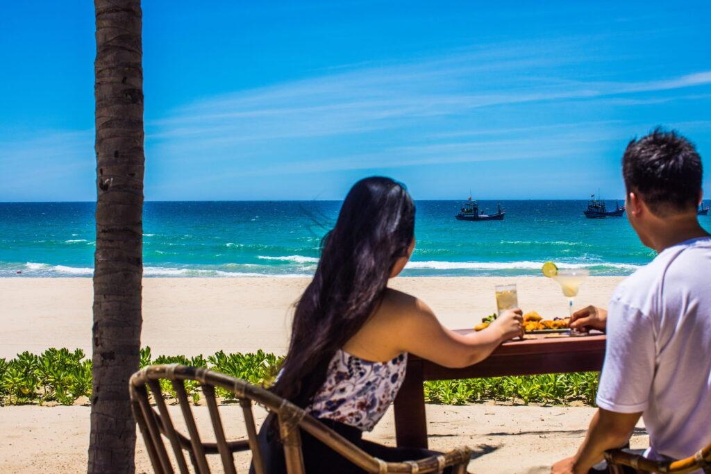 Zwei Leute, die an einem Holztisch am Strand sitzen, mit Getränken und Snacks, den Blick auf das Meer gerichtet, im Hintergrund sind Boote zu sehen