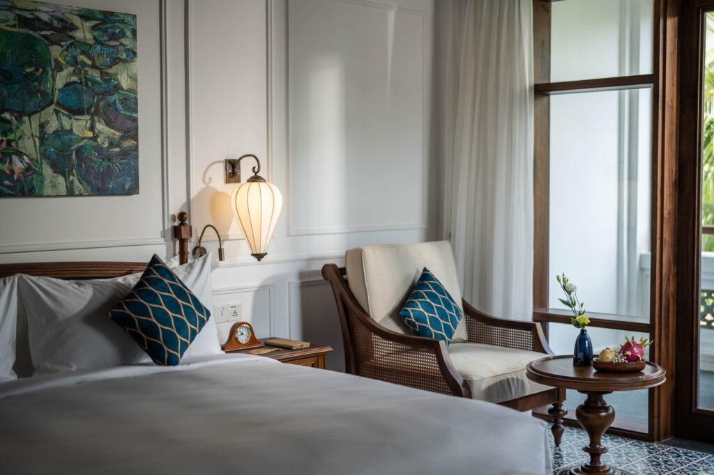 Eine Ecke im Schlafzimmer mit einem Bett, einem Holzsessel mit einem blau gemusterten Kissen, einem kleinen runden Holztisch mit einer Vase und Blumen, einer Wandlampe und einem großen Fenster mit Vorhängen