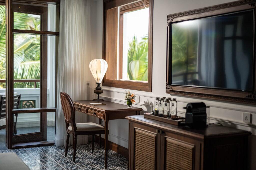 Eine Ecke im Hotelzimmer im „The Anam Mui Ne“ mit einem Schreibtisch und einem Stuhl aus Holz am Fenster, einem an der Wand montierten Flachbildfernseher und einem Schrank mit Kaffeemaschine und Glasflaschen
