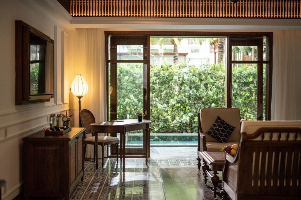 Raumansicht mit Holzmöbeln, darunter ein Stuhl, ein Tisch und ein Sofa, die auf eine Glastür blicken, die zu einem privaten, von Grün umgebenen Pool im luxuriösen Resort „The Anam Mui Ne“ führt