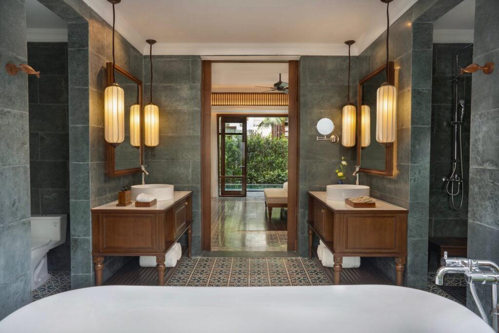Das Badezimmer im luxuriösen Resort „The Anam Mui Ne“ mit zwei Waschtischen aus Holz, rechteckigen Spiegeln, Wandleuchten und einer Badewanne im Vordergrund, von der aus man in einen Raum mit Glastüren gelangt, die zum Garten führen