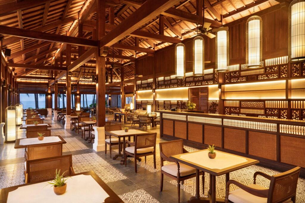 Geräumiges Open-Air-Restaurant mit Holzbalken, Stühlen aus geflochtenem Rattan, quadratischen Tischen mit kleinen Pflanzen und hängenden Laternenleuchten im luxuriösen Resort „The Anam Mui Ne“