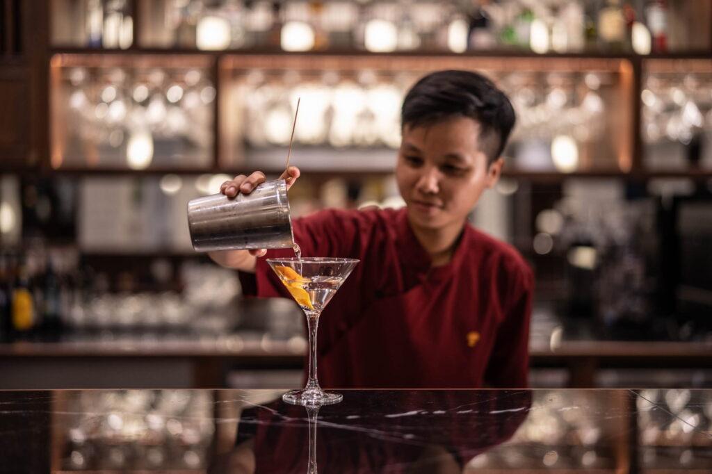 Ein Barkeeper in einem roten Hemd, der hinter der Theke einen Cocktail aus einem Shaker in ein Martini-Glas gießt