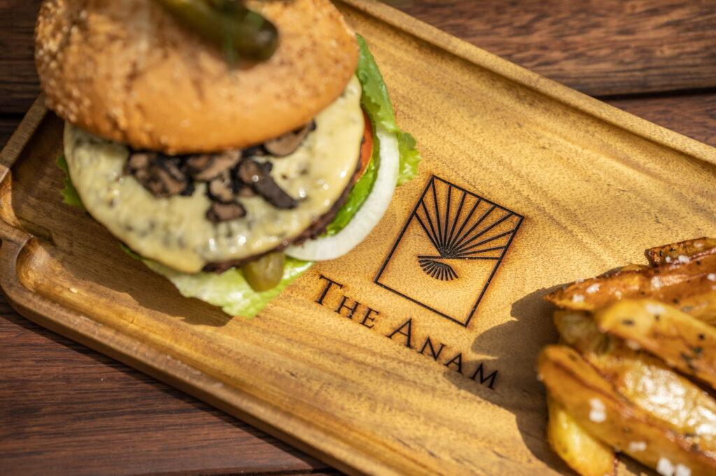 Burger mit Salat, Tomate, Käse und einer Gewürzgurke auf einem Holztablett, in das das „THE ANAM“-Logo eingraviert ist, daneben eine Portion Pommes