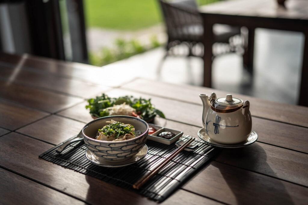 Tischdekoration mit einer Schüssel voller Essen, garniert mit Grünzeug, einem kleinen Teller, Essstäbchen und einer Keramik-Teekanne auf einem Holztisch, im Hintergrund sind verschwommene Stühle und Grünpflanzen zu sehen