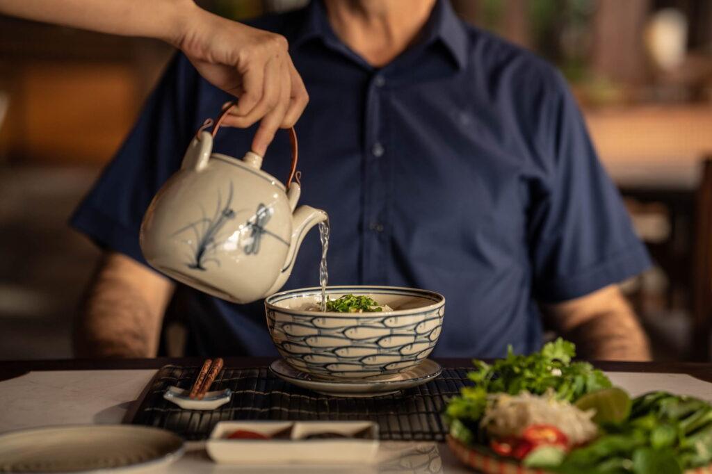 Eine Person in einem blauen Hemd gießt Brühe aus einer Keramikkanne in eine gemusterte Schüssel auf einem Tisch, auf dem Essstäbchen und Teller mit Essen stehen – im Restaurant „The Anam Mui Ne“
