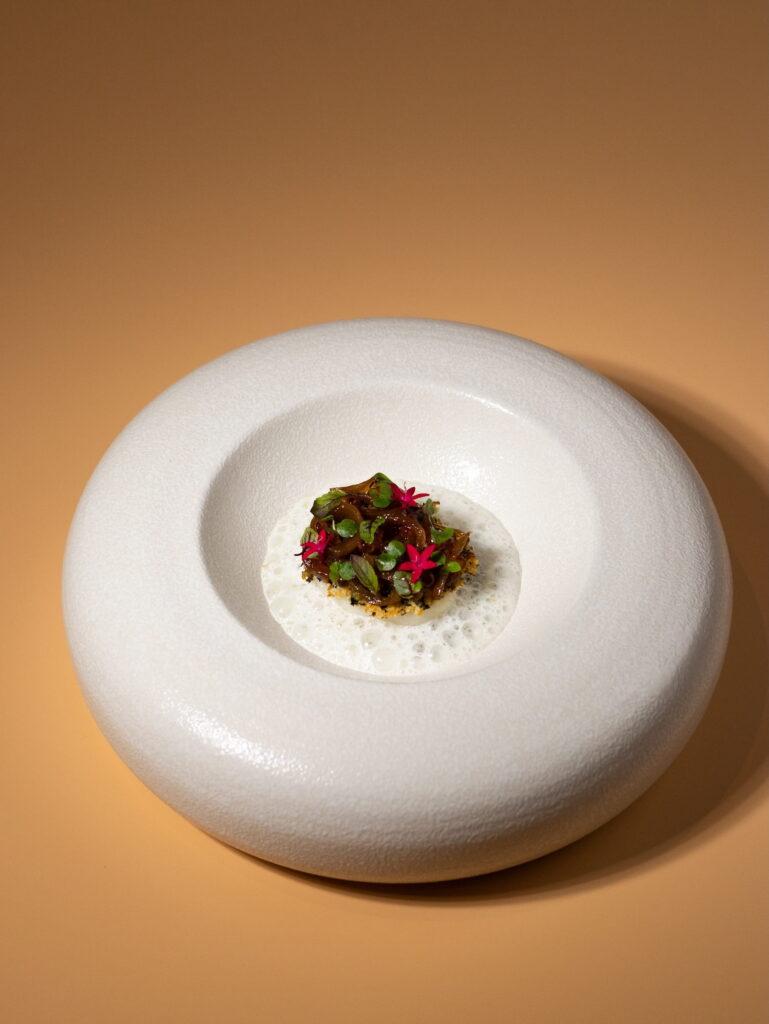 Ein kleines, kunstvoll angerichtetes Gericht mit essbaren Blüten und grünen Kräutern auf einem weißen, runden, flachen Teller vor einem beigen Hintergrund