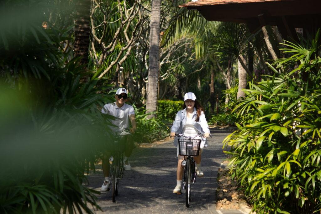 Zwei Personen fahren mit dem Fahrrad auf einem von Palmen und tropischen Pflanzen gesäumten Weg im The Anam Cam Ranh Resort.