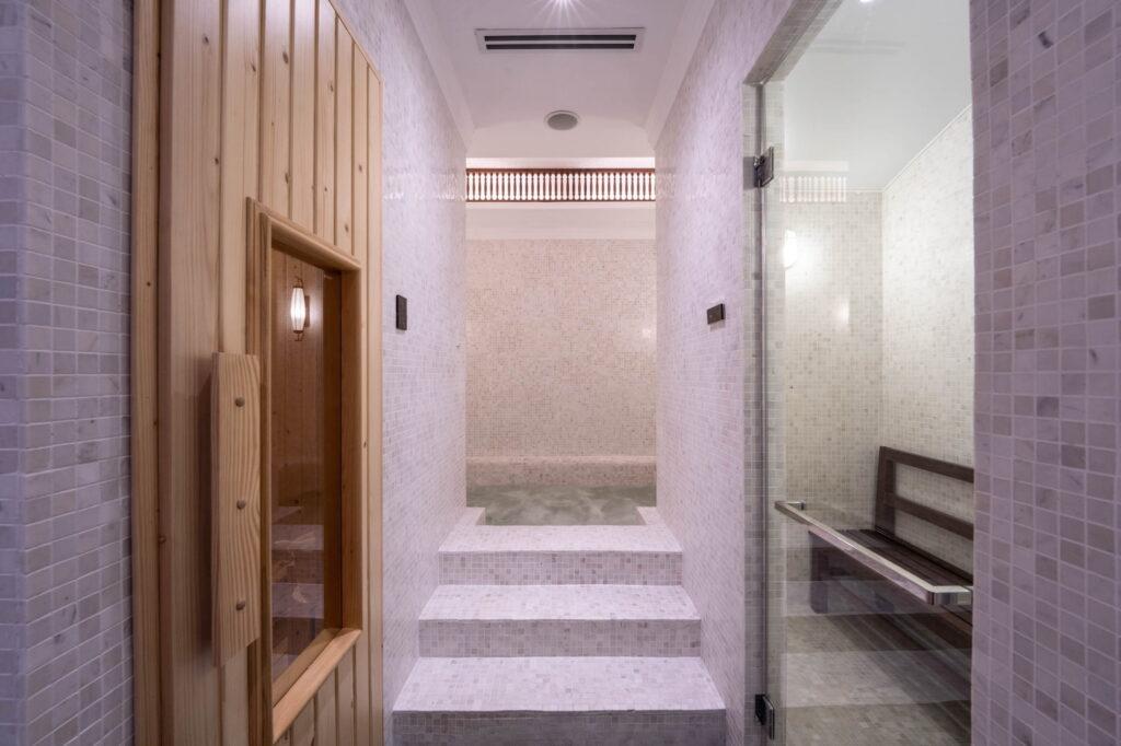 Wellnessbereich im Innenbereich mit gefliesten Wänden, einer Saunatür aus Holz auf der linken Seite, Stufen, die zu einem kleinen Becken führen, und einer Glastür auf der rechten Seite, hinter der eine Bank zu sehen ist