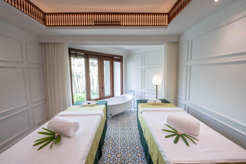 Ein Raum im „The Anam Spa“ mit zwei Massagetischen, die mit weißen Laken und grünen Läufern bedeckt sind – auf jedem liegt ein aufgerolltes Handtuch und ein grünes Blatt –, einer freistehenden Badewanne neben Fenstern mit Holzrahmen und einer Stehlampe in einem Raum mit Fliesenboden