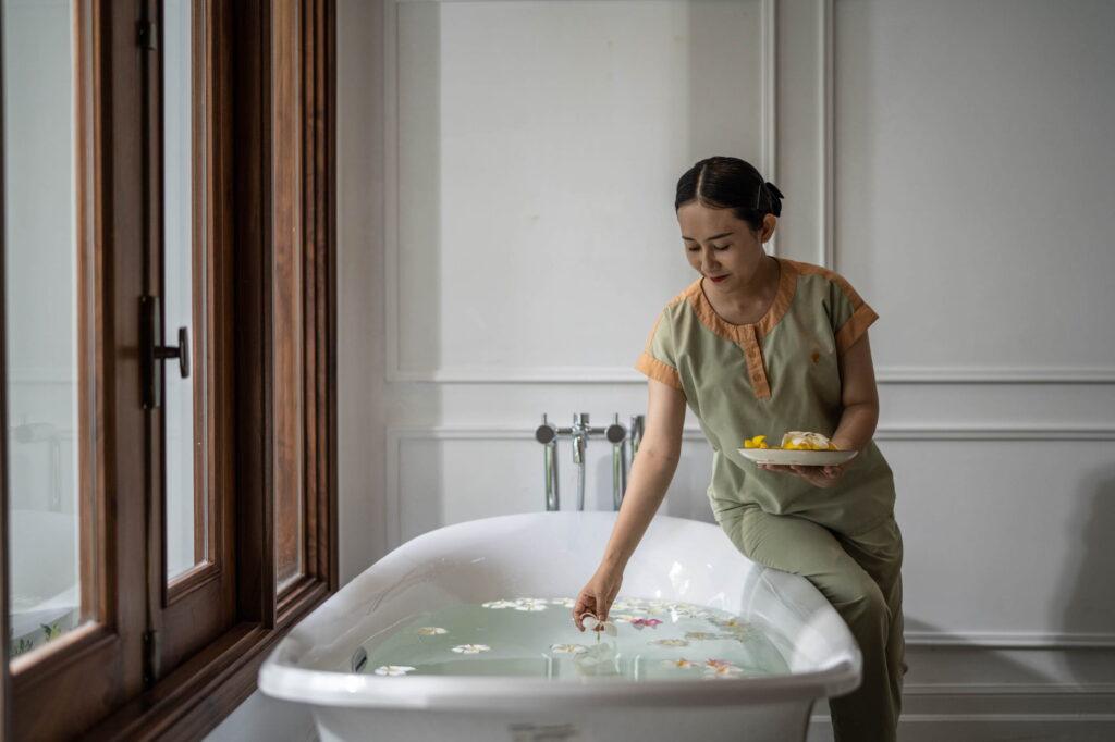 Eine Frau in grüner Kleidung sitzt am Rand einer weißen Badewanne, die mit Wasser und schwimmenden Blumen gefüllt ist, und hält einen Teller mit gelben Blumen in der Hand – in einem Raum mit weißen Wänden und Holzfensterrahmen im The Anam Spa