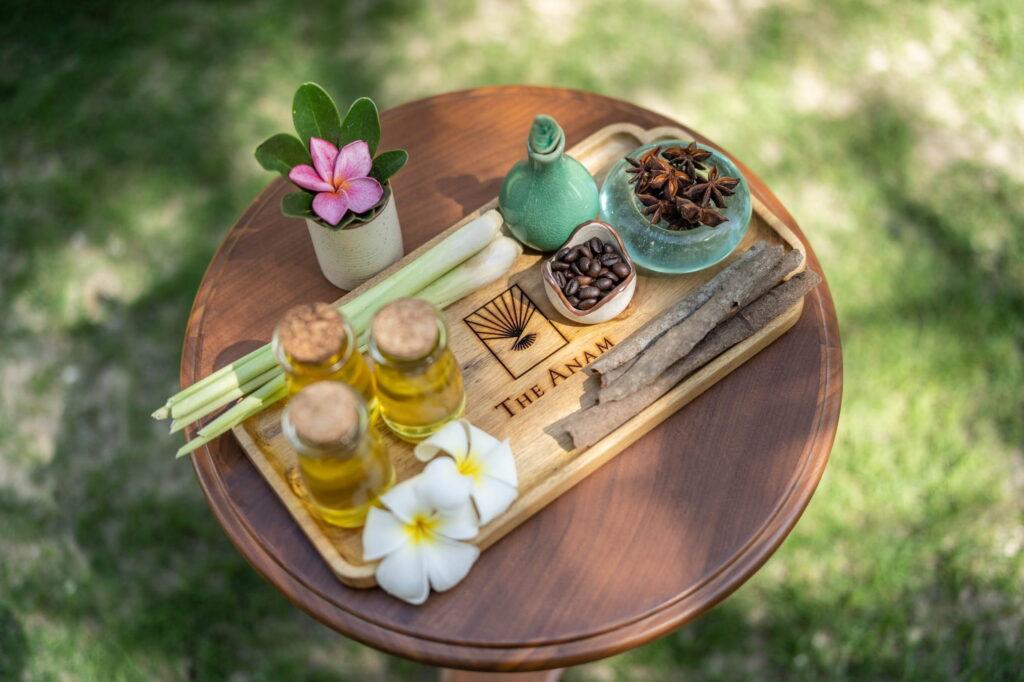 Ein Holztablett auf einem runden Tisch, darauf kleine Ölfläschchen, Kaffeebohnen, Sternanis, Zitronengrasstängel, zwei weiße Blumen, eine rosa Blume in einem kleinen Topf und ein Holzbrett mit der Gravur „THE ANAM“