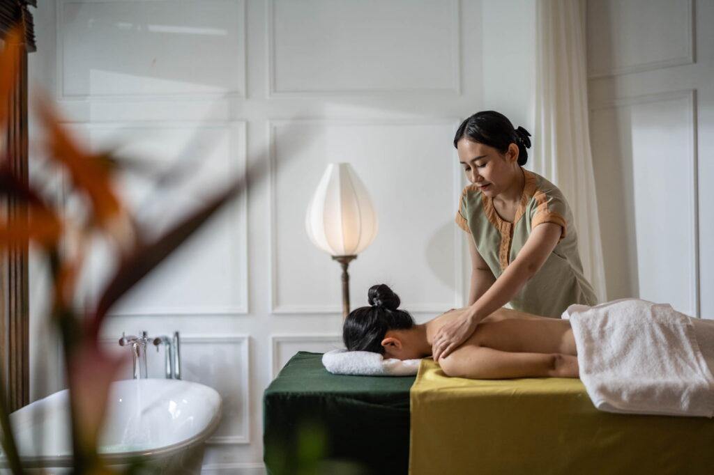 Eine Frau, die auf einer Behandlungsliege in einem Spa-Raum mit weiß getäfelten Wänden und einer Stehlampe im „The Anam Spa“ eine Rückenmassage bekommt