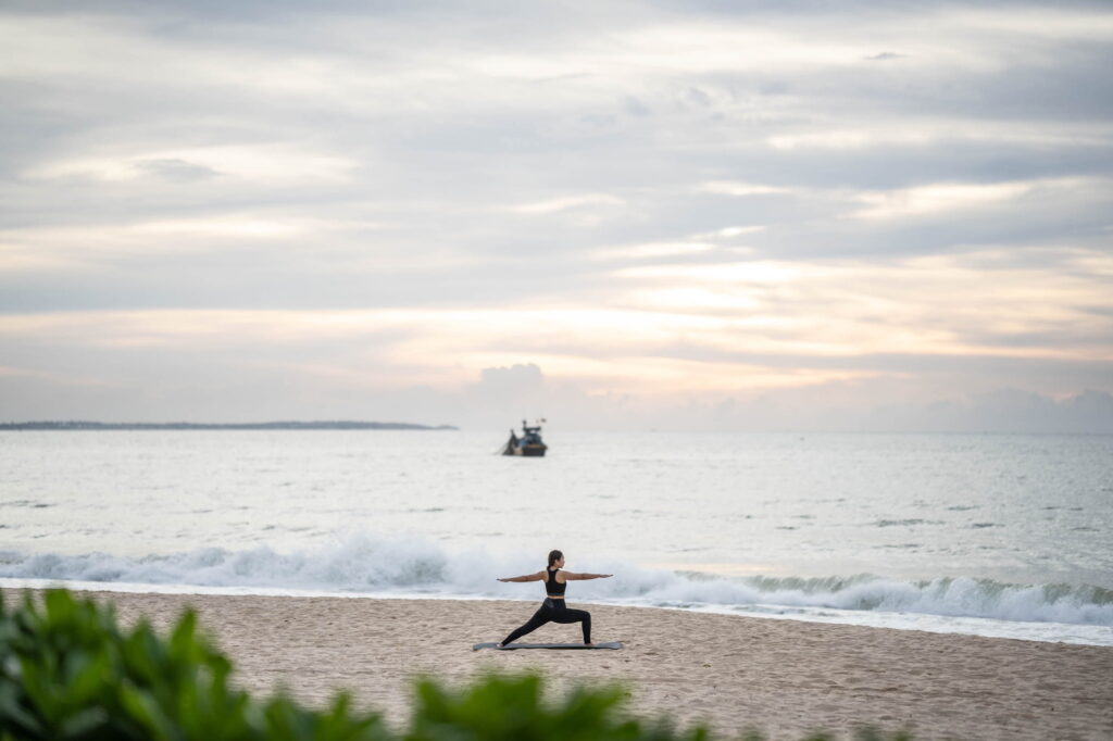 Jemand, der an einem Sandstrand in der Kriegerpose Yoga macht, mit Meereswellen und einem Boot in der Ferne