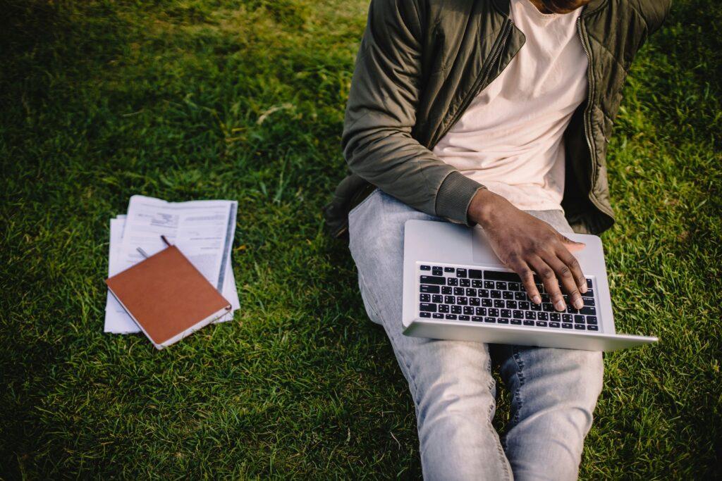 Person sitzt auf Gras und tippt auf einem Laptop, neben ihr liegen ein Notizbuch und Papiere