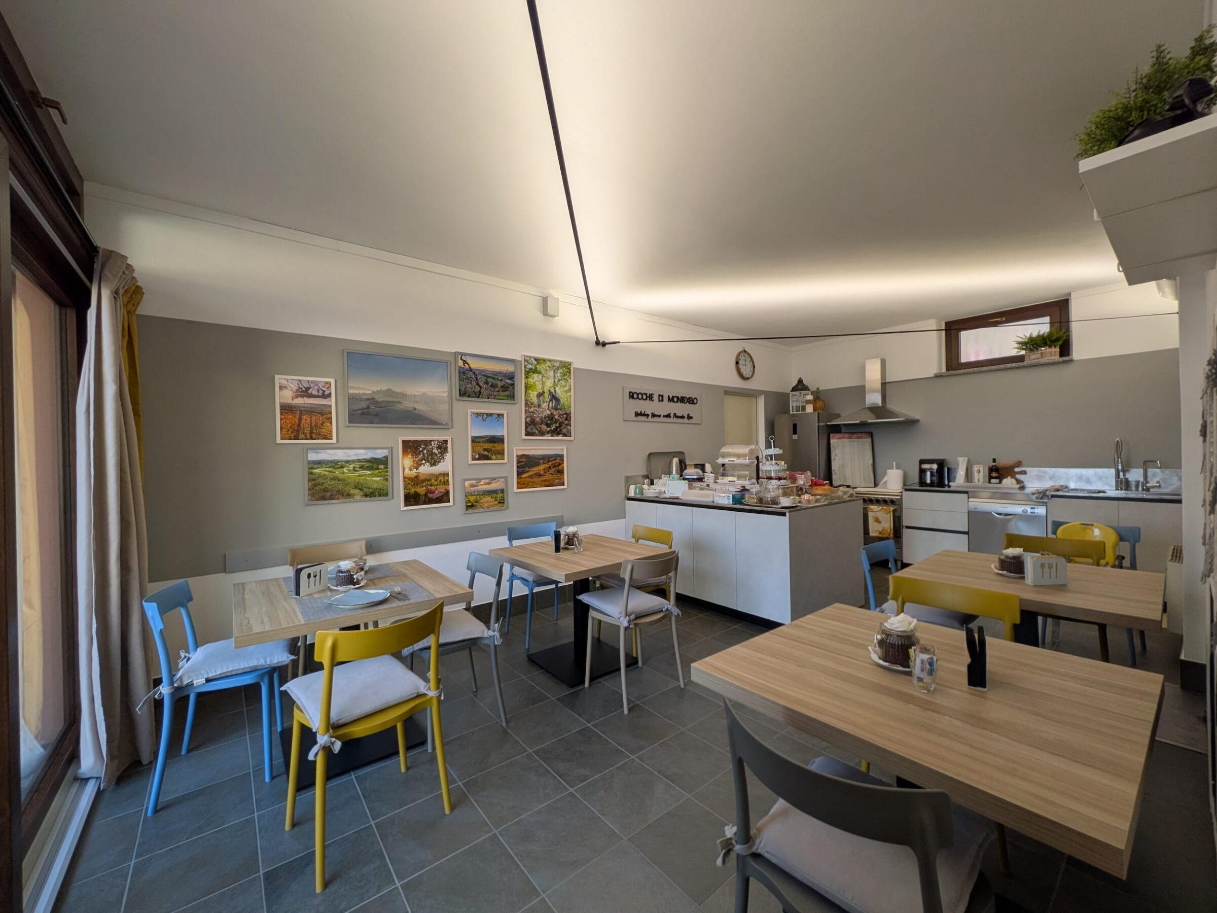 Der Frühstücksraum in den Ferienwohnungen „Rocche di Montexelo“ mit mehreren Tischen und bunten Stühlen, einer mit Landschaftsgemälden geschmückten Wand und einer Buffetinsel in der Mitte