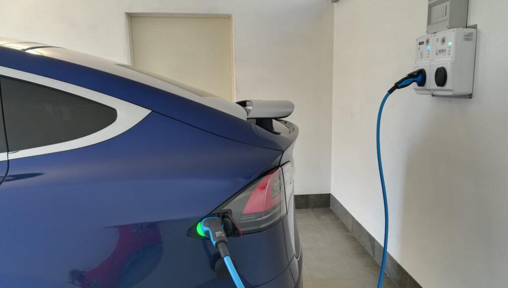 Das Heck eines blauen Elektroautos, das an einer Wandladestation in einem Innenraum mit grauem Boden und weißen Wänden in Rocche di Montexelo aufgeladen wird