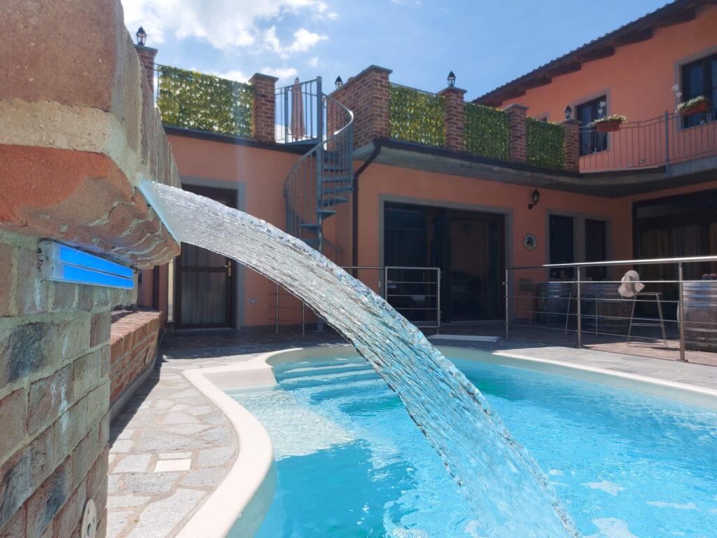 Pool mit einem aus Mauerwerk gestalteten Wasserfall vor den Ferienwohnungen „Rocche di Montexelo“, mit Balkon und Geländer