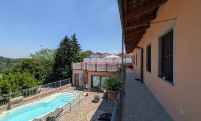 Blick vom Balkon auf den Pool mit Liegestühlen und Sonnenschirmen neben dem hellen Gebäude mit Holzdach in Rocche di Montexelo, im Hintergrund die grüne Landschaft des Piemonts