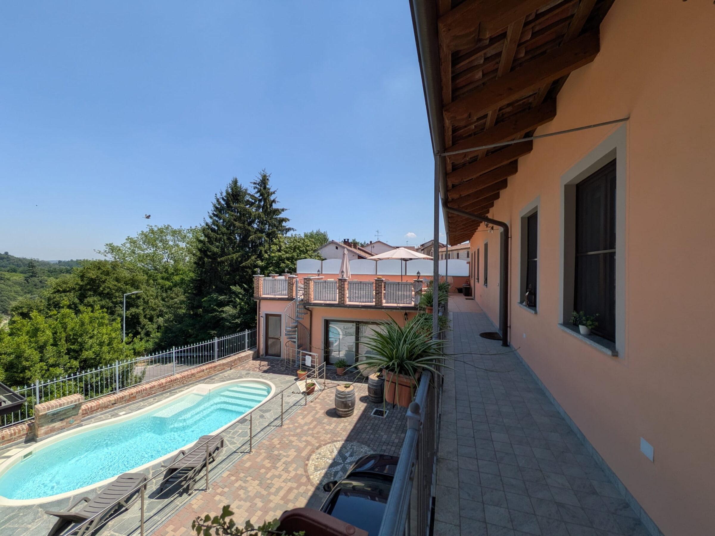 Blick vom Balkon auf den Pool mit Liegestühlen und Sonnenschirmen neben dem hellen Gebäude mit Holzdach in Rocche di Montexelo, im Hintergrund die grüne Landschaft des Piemonts