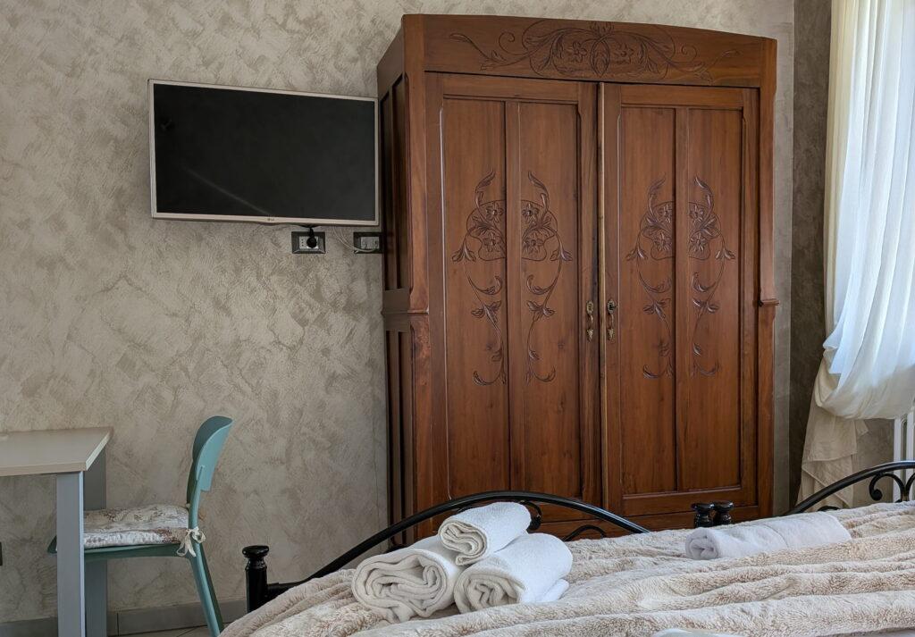 Ein Schlafzimmer mit einem geschnitzten Holzschrank, einem an der Wand montierten Fernseher, einem kleinen Schreibtisch mit Stuhl und einem Bett, vor dem aufgerollte Handtücher liegen, vor einem Fenster mit einem weißen, luftigen Vorhang in einer Wohnung in Rocche di Montexelo