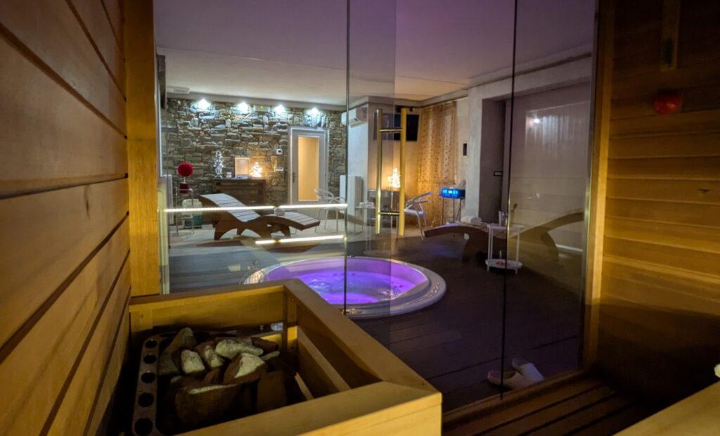 Blick durch eine Glaswand im Wellnessbereich von Rocche di Montexelo, im Vordergrund zu sehen sind ein Whirlpool, Liegestühle und ein Saunaofen mit Steinen