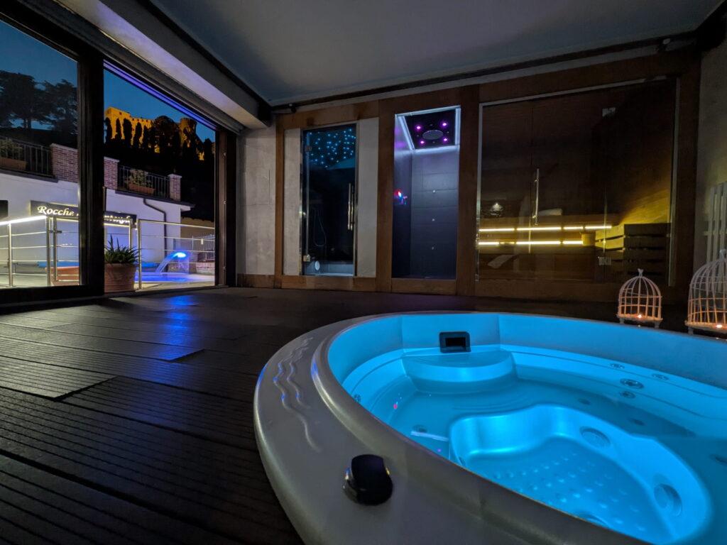 Der Innenbereich des Wellnessbereichs „Rocche di Montexelo“ mit einem beleuchteten Whirlpool, raumhohen Fenstern und einem Blick auf eine Terrasse mit Sitzgelegenheiten bei Nacht