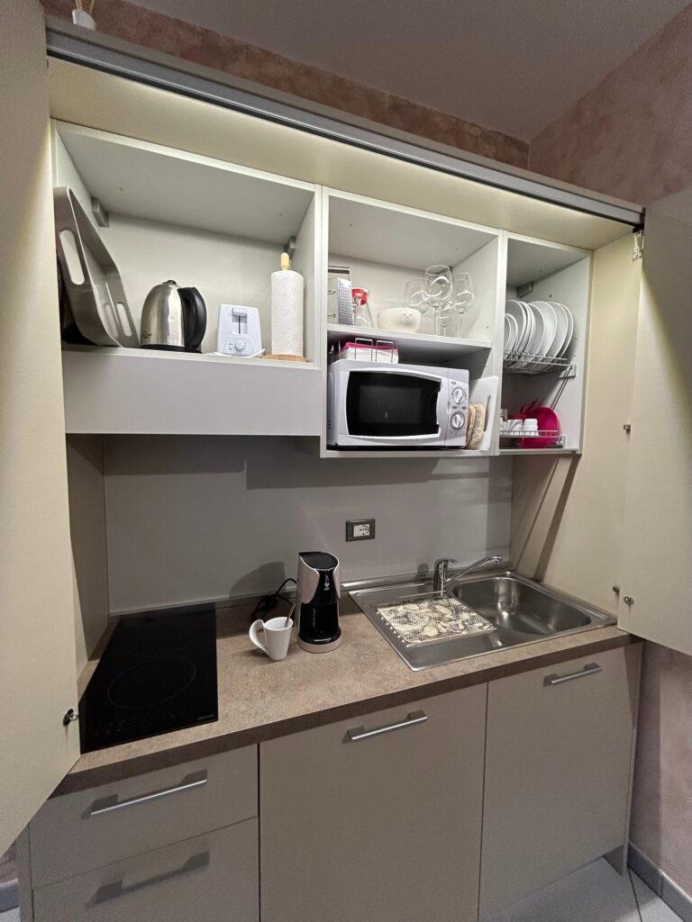 Kleine Kochnische in einer Ferienwohnung in Rocche di Montexelo, ausgestattet mit Spüle, Kochfeld, Kaffeemaschine, Mikrowelle, Wasserkocher, Geschirr und Küchenutensilien, die in offenen und geschlossenen Schränken aufbewahrt werden.