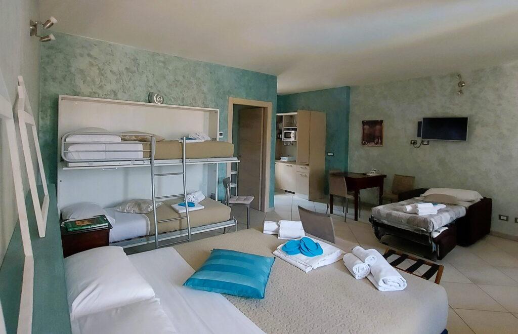 Schlafzimmer in den Ferienwohnungen „Rocche di Montexelo“ mit einem Etagenbett, einem Einzelbett, einem Doppelbett, einem Schreibtisch, einem an der Wand montierten Fernseher und türkisfarbenen Wänden.