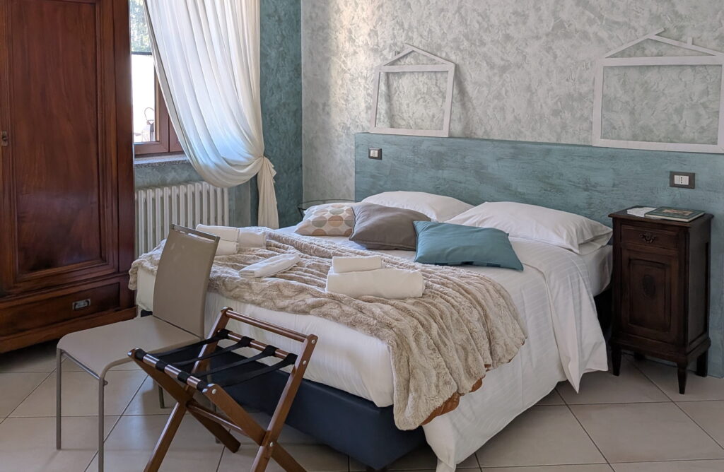 Schlafzimmer in den Ferienwohnungen „Rocche di Montexelo“ mit einem Doppelbett, zwei Nachttischen, einem Stuhl, einem Kofferablage und einem Fenster mit einem weißen Vorhang