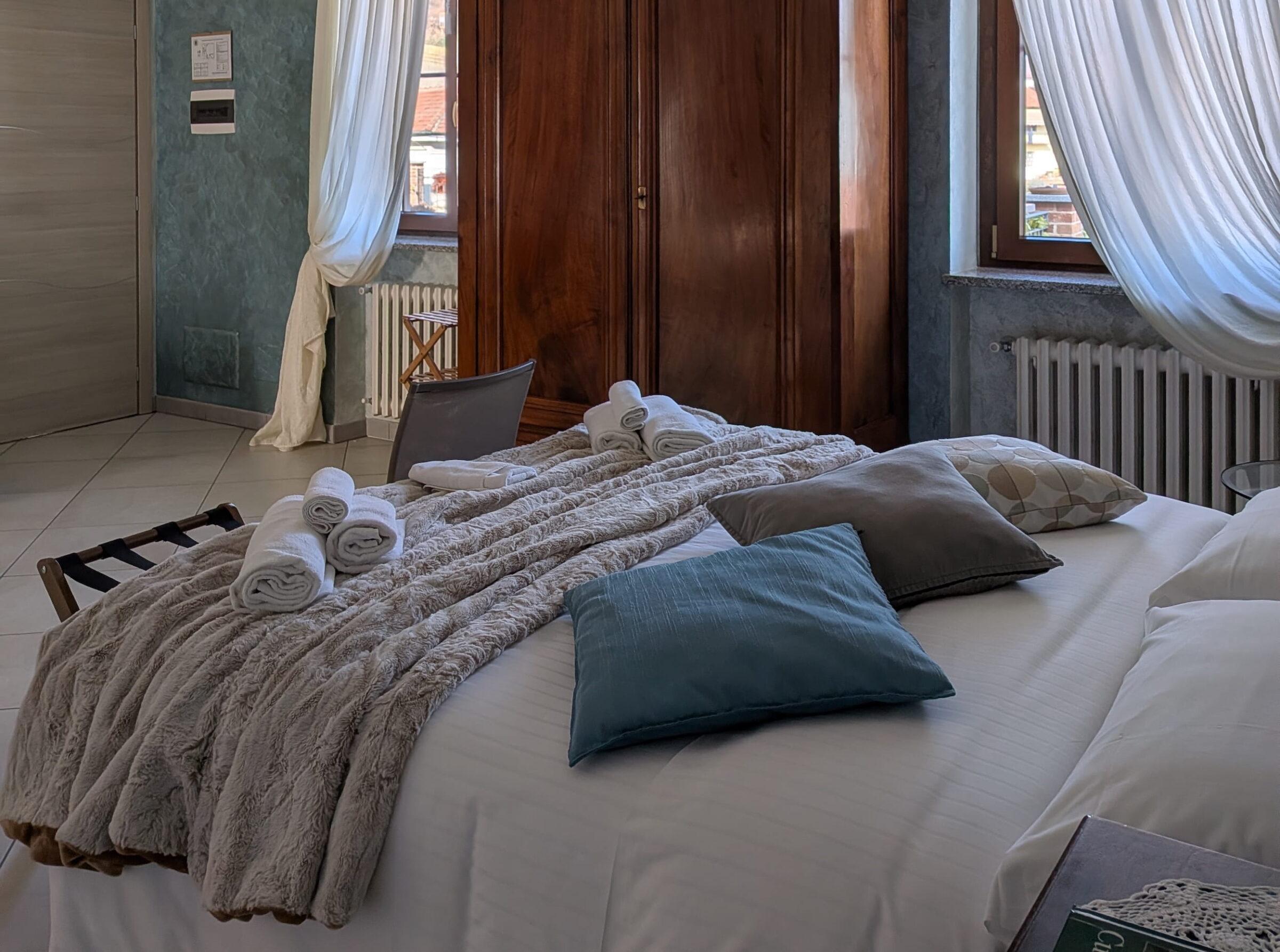 Schlafzimmer in den Ferienwohnungen „Rocche di Montexelo“ mit einem großen Bett, weichen Kissen, einer flauschigen Decke, aufgerollten Handtüchern und zwei Fenstern mit weißen Vorhängen.