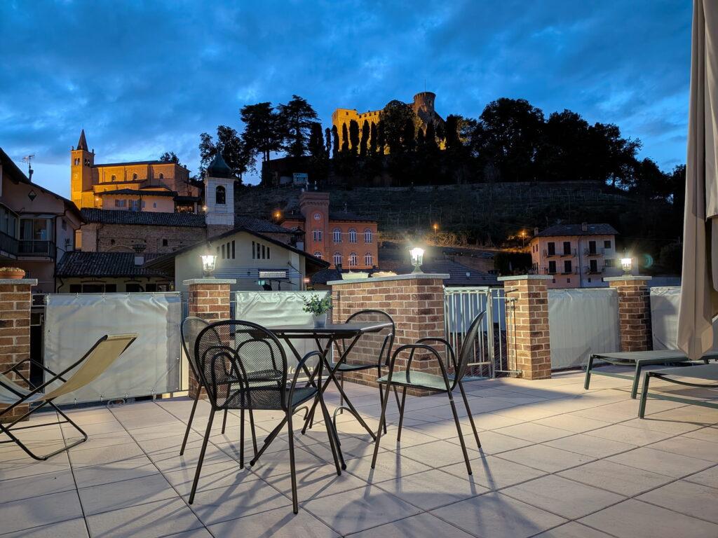 Die Terrasse von Rocche di Montexelo mit Stühlen und einem Tisch vor der Kulisse der beleuchteten Burg und des Dorfes in der Abenddämmerung
